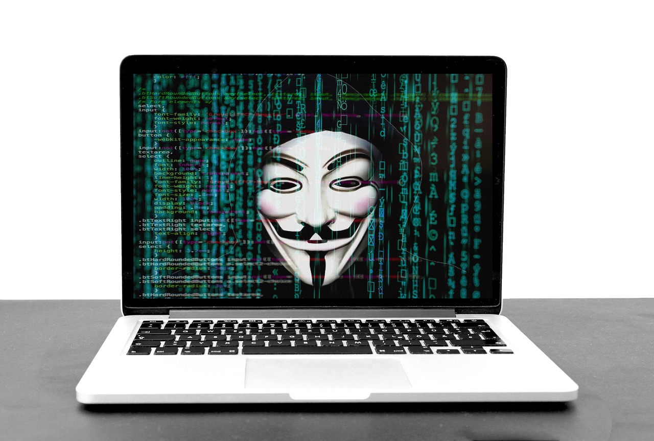 Laptop z wyświetloną maską anonimowego hakera i kodem symbolizujący zagrożenia cybernetyczne